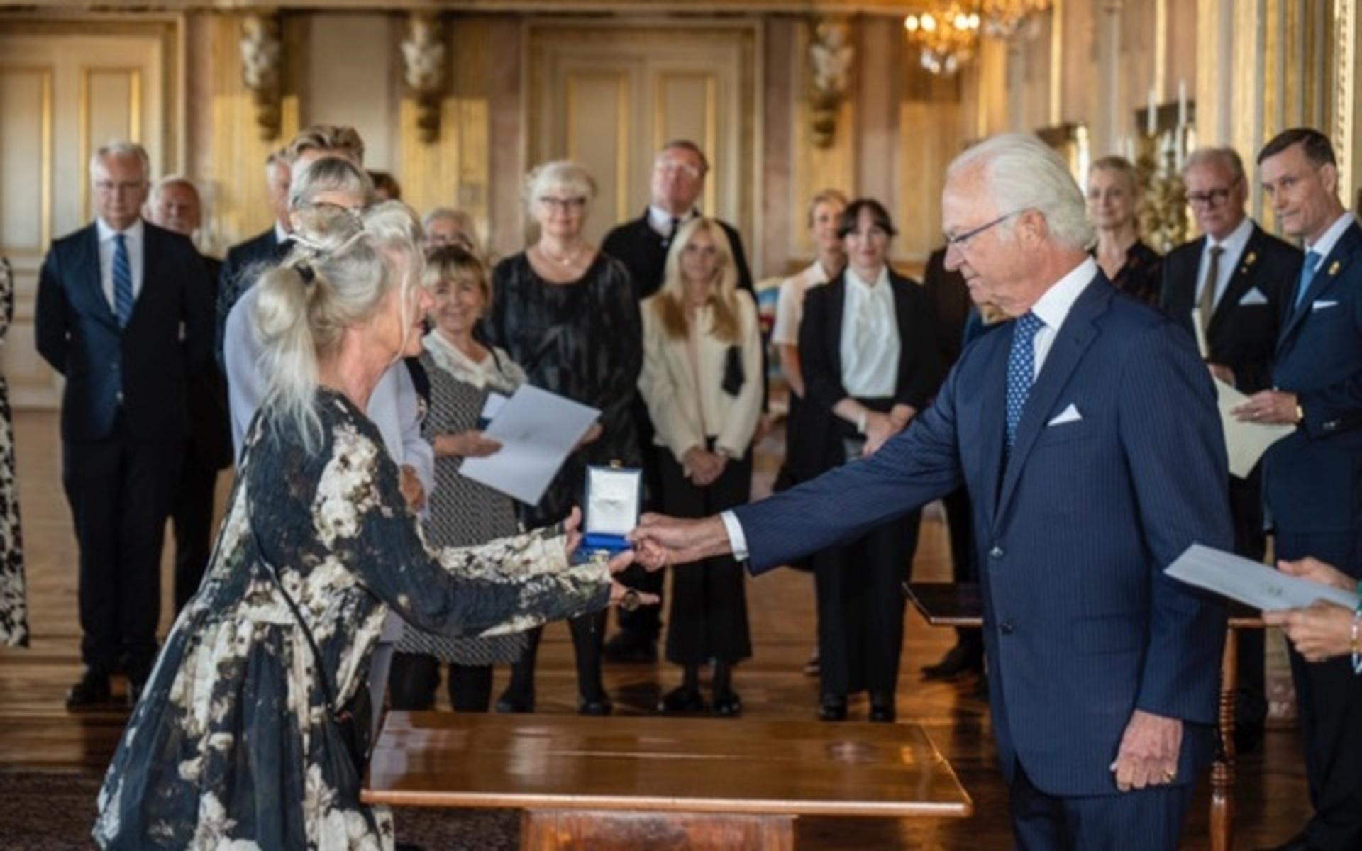 Här räcker kungen över medaljen till Irène Sjöberg-Lundin. I bakgrunden syns bland andra Beatrice Ask (M), tidigare kulturminister, och Per Nuder (S), som var där som anhörig och inte fick ta någon egen medalj med sig hem.